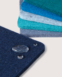 Close-up of a stack of colorful felt squares in blue, teal, and gray by graf lantz. The top German Merino wool piece is dark blue, showcasing water droplets for its water-repellent property—ideal for sustainable home décor aficionados.