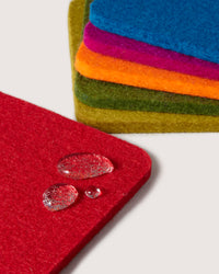 Close-up of vibrant German Merino wool coasters by graf lantz, artistically fanned out on a light background. The top coaster is red with droplets, with green, orange, purple, and blue underneath. This chic and sustainable home décor adds both style and eco-friendliness.