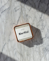 On a marble surface, square Merino wool coasters labeled Bierfilzl rest in a dish. The sunlight casts shadows, emphasizing their handcrafted quality by graf lantz in Los Angeles.