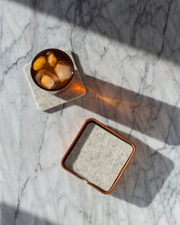 A dark drink with ice rests on a square Merino wool coaster from graf lantz on a marble surface, beside its empty pair. Sunlight casts long shadows, enhancing the elegance of this Los Angeles-crafted scene.