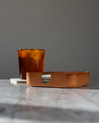 A glass of iced tea rests on a coaster beside a rectangular graf lantz Kobon leather tray holding round coasters, set on a marble surface against a gray background.