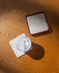 A glass of ice water rests on a light-colored square graf lantz coaster on a wooden table, handcrafted in Los Angeles, with a matching holder nearby. Sunlit shadows cast patterns across the surface.