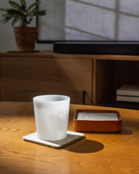 A white mug rests on a Merino wool coaster atop a wooden table, next to a brown ceramic container. Sunlight streams through the window, casting shadows. In the background, a plant in a pot sits alongside a Kobon leather tray by graf lantz, handcrafted in Los Angeles.