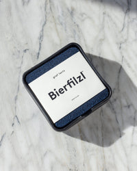 A black square container on a marble surface holds a stack of soft Merino wool blue Bierfilzl coasters by graf lantz. The top coasters label reads Bierfilzl and graf lantz, indicating its made in the USA. Shadows add depth to the marble scene.