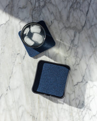 A glass of iced water rests on a dark square Bierfilzl coaster from graf lantz on a light marble surface. Its Merino wool charm enhances the scene, while another coaster lies nearby, with sunlight casting soft shadows across the marble.