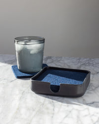 A glass of ice water rests on a blue Bierfilzl coaster from graf lantz on a marble surface. Beside it, an open Kobon leather tray from graf lantz holds additional blue coasters. Soft natural light illuminates the scene, casting gentle shadows.