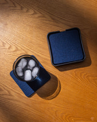 A glass of iced drink sits on a dark blue Graf Lantz Bierfilzl coaster, with a stack of matching coasters nearby on the wooden surface. Soft lighting casts warm shadows that accentuate the wood grain.