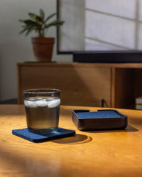A glass of iced water rests on a blue Bierfilzl coaster from graf lantz on a wooden table, beside an empty Kobon leather tray. Sunlight casts shadows over the scene, with a potted plant and TV screen softly blending into the blurred background.