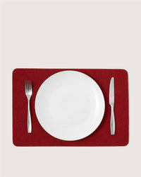 A white empty plate is centered on a red Graf Lantz placemat, with a fork on the left and a knife on the right, neatly arranged on a light-colored surface.