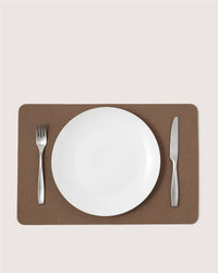 A white plate sits centered on a brown Graf Lantz Merino wool placemat, with a fork to the left and a knife to the right, creating a simple and stylish table setting against a light background.
