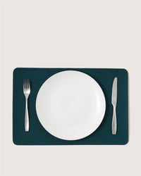 A white plate is centered on a Graf Lantz dark green rectangular placemat, with a silver fork on the left and a silver knife on the right, all placed on a light-colored surface.
