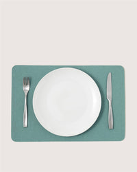 A white empty plate sits on a green Graf Lantz Merino wool placemat, with a fork to the left and knife to the right, creating an elegant designer table setting against a plain, light background.