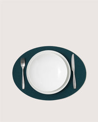 A white Graf Lantz plate and bowl sit on a dark green oval placemat, with a silver fork to the left and a silver knife to the right, all arranged on a light background.