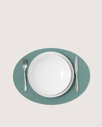 A white bowl on a white plate rests on a green oval Graf Lantz Merino wool placemat, with a fork to the left and knife to the right, all neatly arranged on a light background.