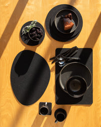 Overhead view of a wooden table featuring graf lantz wool coasters with black plates, a bowl, and mugs. Plates hold small cakes and a dish filled with blackberries and sage, while sunlight casts shadows across the scene.