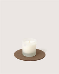 A white candle in a frosted glass holder rests on a round, sustainable Merino wool trivet from Graf Lantz against a plain light background.
