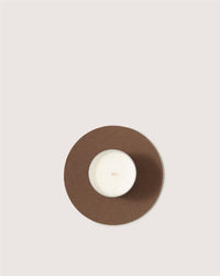 A white candle in a glass holder sits on a round brown Merino wool trivet by Graf Lantz, viewed from above against a plain light background—a stylish and sustainable kitchen accessory.