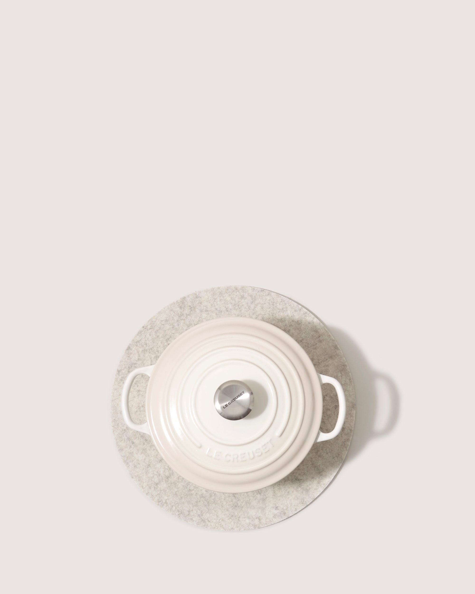 A stylish white casserole dish, heat-resistant and featuring a silver knob, rests on a gray circular mat. Viewed from above, it shows two side handles and the graf lantz brand embossed on the lid against a plain light background.