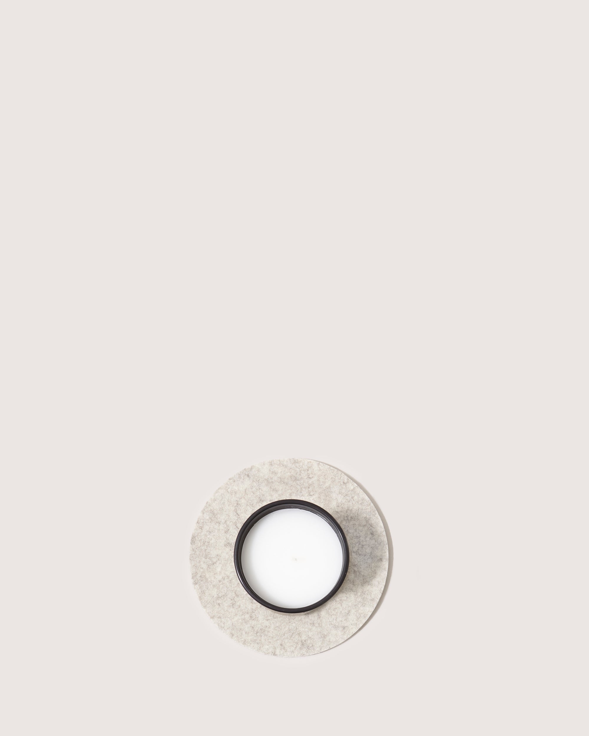 A minimalist photo features a single black candle on a circular white felt mat, designed as a trivet by graf lantz, set against a plain light background.