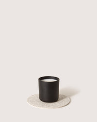 A black candle in a minimalist container rests on a round, light gray Merino wool coaster by graf lantz. The plain, neutral background highlights the simple elegance of the Oeko-Tex® Certified setup.