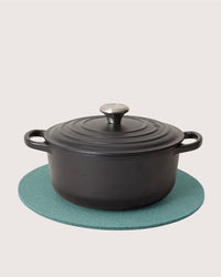 A black cast iron Dutch oven with lid and handles sits on a round, heat-resistant Merino wool trivet from Graf Lantz, set against a plain light background.