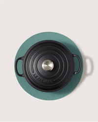 A black Le Creuset cast iron Dutch oven with a silver knob rests on a round green Graf Lantz Merino wool trivet, viewed from above against a light background. The sustainable pad is both stylish and heat-resistant.