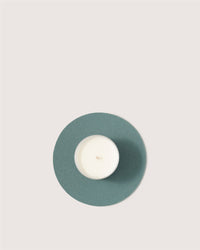 A small white tealight candle in a metal holder sits on a round, teal Merino wool trivet by Graf Lantz—a sustainable kitchen accessory—set against a plain, light background.
