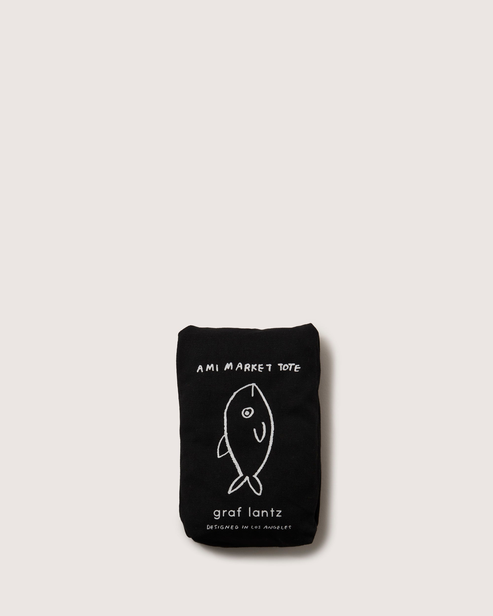 A folded black fabric tote bag by graf lantz, showcasing a simple white fish drawing reminiscent of Japanese fish markets, features AMI MARKET TOTE text on a plain white backdrop.