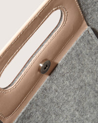 The graf lantz Asobi Bag is a minimalist clutch made from gray Merino wool, with a rectangular cut-out handle reinforced by a beige strap. The lower half features small, evenly spaced holes, all set against a light neutral background for an elegant and simple look.