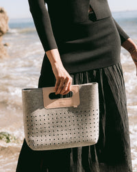 The graf lantz Asobi Bag is a minimalist clutch made from gray Merino wool, with a rectangular cut-out handle reinforced by a beige strap. The lower half features small, evenly spaced holes, all set against a light neutral background for an elegant and simple look.