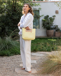 A woman stands barefoot on a stone path in a garden, wearing a white shirt and pants. Shes holding a yellow perforated Graf Lantz Asobi Bag and looking at the camera. Bushes and a white building with a window are in the background, enhancing the serene ambiance.