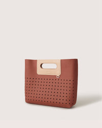 The Graf Lantz Asobi Bag is a chic brown handbag with a perforated lower half, featuring a minimalist tan handle cutout for easy carrying. A small metal ring accent adds charm near the handle, and the plain white backdrop enhances its sleek elegance.