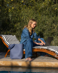 A person in denim relaxes on a striped lounge chair by the pool, working on a laptop. A Graf Lantz sustainable Merino Wool Tote lies beside them, highlighting its everyday functionality. The scene is framed by lush greenery in the background.
