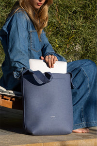 A woman in a denim outfit enjoys the blend of style and function outdoors as she places a white laptop into her sustainable blue Graf Lantz tote bag, surrounded by greenery and sunlight.