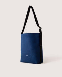 A graf lantz blue Merino Wool tote bag with a black adjustable shoulder strap against a plain light background. The water-resistant design includes a small white logo text near the bottom.