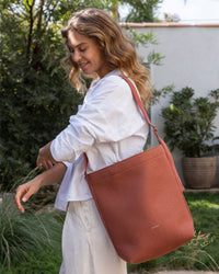 Standing outdoors amidst lush greenery, a person with wavy hair and a smile looks down while wearing a white shirt. Over their shoulder is Graf Lantzs large terracotta Merino Wool Tote, blending sustainable style with everyday functionality.