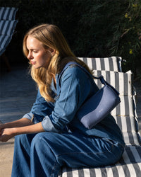 With long blonde hair, a person relaxes on a striped lounge chair, showcasing a minimalist style in denim and holding a Graf Lantz Campus Shoulder Bag. Outdoors on a sunny day, surrounded by greenery, they exude relaxation.