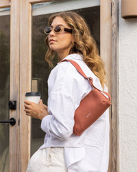 A wavy-haired individual in sunglasses, a white Merino Wool shirt, and cream pants holds a reusable coffee cup. They carry Graf Lantzs rust-colored Campus Shoulder Bag and stand before a glass door.