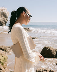 A person in a white dress and sunglasses stands on a rocky beach, holding a sustainable Deco Edge Pouch by Graf Lantz. Ocean waves gently hit the shore under a clear blue sky.