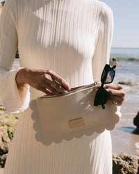 A person in a Graf Lantz long-sleeve, ribbed Merino wool cream dress stands on the beach, holding sunglasses and a sustainable Deco Edge Pouch. The ocean and rocks are visible under the clear blue sky.