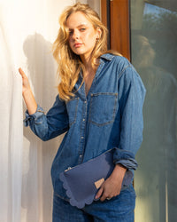 A person with long blonde hair stands by a window, wearing a blue denim shirt and pants while holding a versatile Graf Lantz Deco Edge Pouch. Sunlight filters through the curtain, creating a warm ambiance.