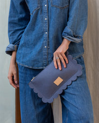 A person in denim holds a Graf Lantz clutch with deco-edge details and a leather patch, their left hand adorned with a silver ring resting on their thigh.