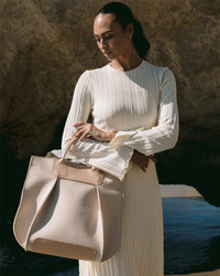 A woman in a white, pleated, long-sleeve dress with a structured silhouette stands by a rocky backdrop holding a large Frankie Tote from Graf Lantz. Sunlight casts soft shadows, highlighting the dresss texture.