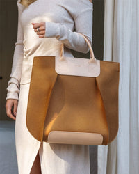 A person in a light beige dress holds the stylish Frankie Tote by Graf Lantz, featuring rounded edges and beige handles as an ideal versatile companion, set against light-colored curtains.