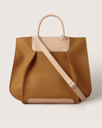 The Graf Lantz Frankie Tote is a chic, large brown bag with beige leather handles and a removable shoulder strap. It features a sleek design, engraved logo on the front, and is set against a light neutral backdrop.