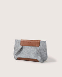 The graf lantz Frankie Clutch, a gray felt pouch with brown leather accents and a rectangular patch, embodies sustainable fashion. Its soft shape highlights Merino wools benefits, standing beautifully against a plain light background.