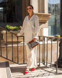 A person in a light-colored suit and sunglasses stands by beige steps, showcasing bright red shoes. They hold an elegant graf lantz Frankie Merino Wool Clutch, emphasizing sustainable fashion benefits as sunlight casts shadows on the ground.