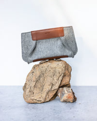 A graf lantz gray felt pouch with a brown leather accent is artfully balanced on stacked rocks against a minimalist concrete background, capturing the essence of sustainable fashion.