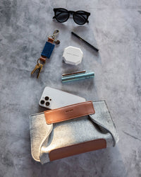 A flat lay of various items on a textured surface: black glasses, a keychain, compact makeup, a pen, wristwatch, smartphone, mascara tube, and a graf lantz Frankie Merino Wool Clutch with gray felt and tan leather accents as a sustainable fashion statement.