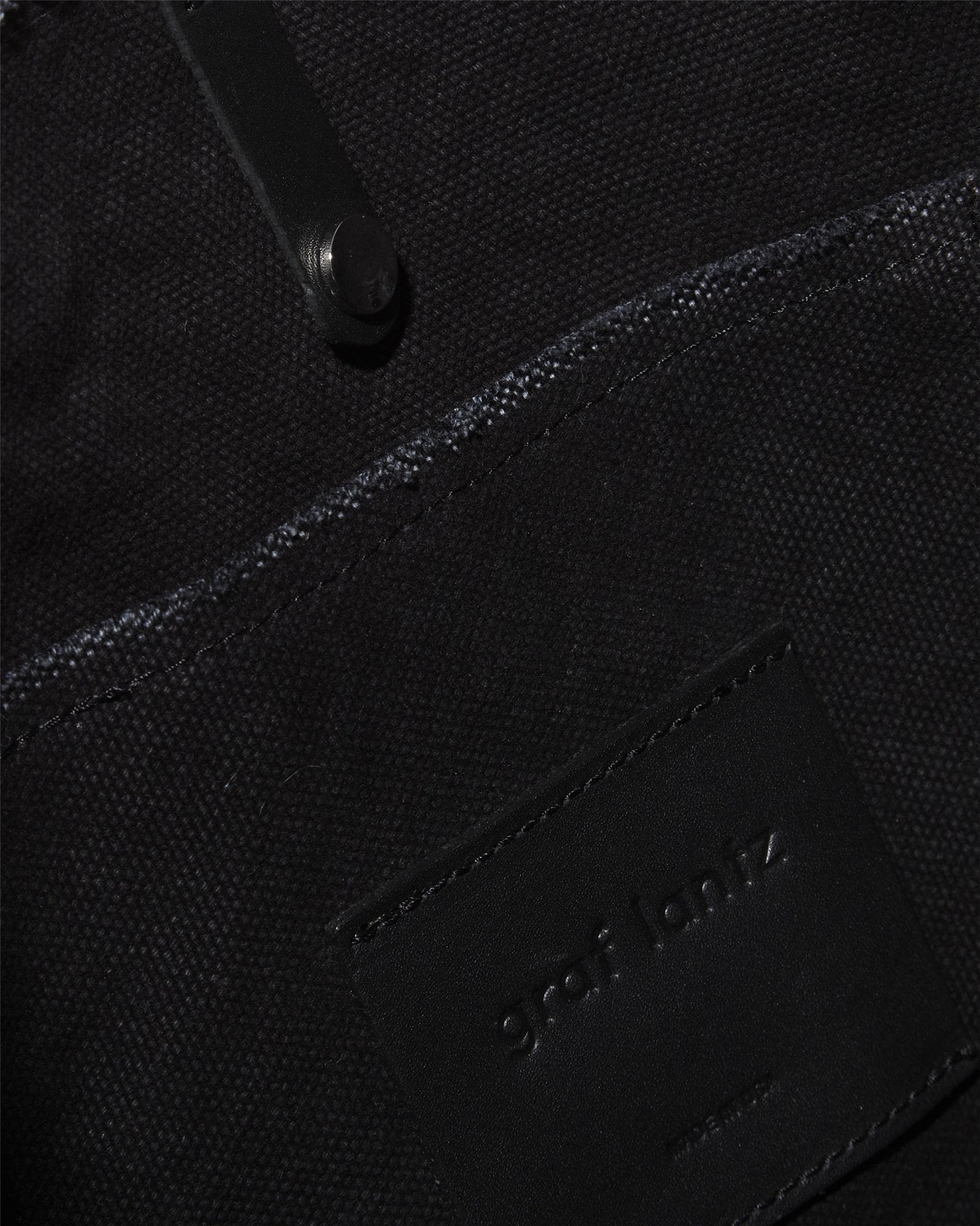 Close-up of a black fabric bag with structured leather handles, snap button, and an embossed Graf Lantz patch. The durable cotton canvas has a textured look, enhancing its chic design.